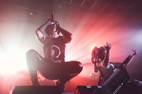 A live music performance featuring a dynamic frontman passionately singing into a microphone, while a female musician plays a keytarnearby. The stage is illuminated with vibrant lights, creating an energetic atmosphere.