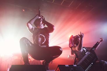 Aesthetic Perfection @ ((szene)) Wien Ein Musiker steht in einer dramatischen Pose mit einem Mikrofon in der Hand auf der Bühne. Neben ihm spielt eine Musikerin eine Keytar. Die Bühne wird von farbigen Lichtern beleuchtet, die eine lebendige und energiegeladene Atmosphäre schaffen.