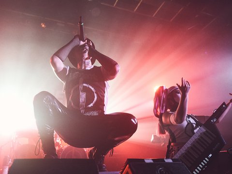 Ein Musiker steht in einer dramatischen Pose mit einem Mikrofon in der Hand auf der Bühne. Neben ihm spielt eine Musikerin eine Keytar. Die Bühne wird von farbigen Lichtern beleuchtet, die eine lebendige und energiegeladene Atmosphäre schaffen.