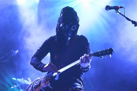 Ein Musiker trägt ein auffälliges Kostüm mit einer Maske, die an einen Vogel erinnert, und spielt Gitarre. Die Beleuchtung im Hintergrund erzeugt eine mystische Atmosphäre mit blauen und violetten Lichtstrahlen.