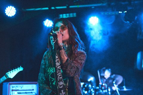 Eine Sängerin mit Sonnenbrille steht auf der Bühne und singt ins Mikrofon. Im Hintergrund ist eine Band zu sehen, die musiziert, während blaue Lichter die Szene beleuchten.