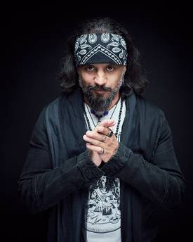 Ein Mann mit langen, dunklen Haaren und einem Bandana schaut ernst in die Kamera. Er trägt ein schwarzes Oberteil und hat die Hände vor der Brust gefaltet. Der Hintergrund ist dunkel, was den Fokus auf sein Gesicht lenkt.