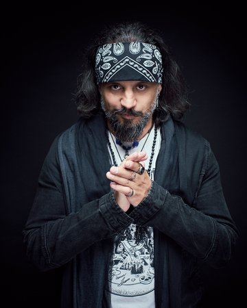 Ein Mann mit langen, dunklen Haaren und einem Bandana schaut ernst in die Kamera. Er trägt ein schwarzes Oberteil und hat die Hände vor der Brust gefaltet. Der Hintergrund ist dunkel, was den Fokus auf sein Gesicht lenkt.