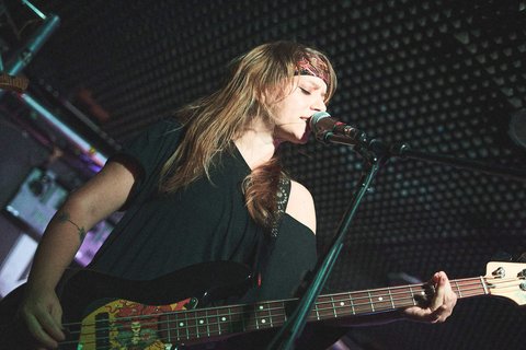 Eine junge Frau spielt Bass und singt in ein Mikrofon. Sie trägt ein schwarzes Shirt und hat lange, lockige Haare. Im Hintergrund ist eine dunkle, strukturelle Wand zu sehen. Die Stimmung der Szene ist musikalisch und intensiv.