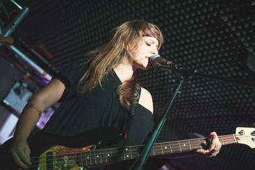 Eine junge Frau spielt Bass und singt in ein Mikrofon. Sie trägt ein schwarzes Shirt und hat lange, lockige Haare. Im Hintergrund ist eine dunkle, strukturelle Wand zu sehen. Die Stimmung der Szene ist musikalisch und intensiv.