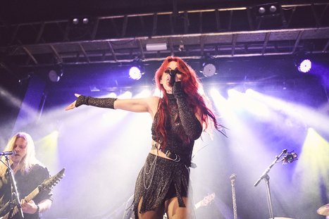 A female singer with long red hair performs on stage, wearing a black outfit with fringe. She gestures passionately while singing into a microphone, surrounded by colorful stage lights and musicians in the background. The atmosphere is energetic and engaging.