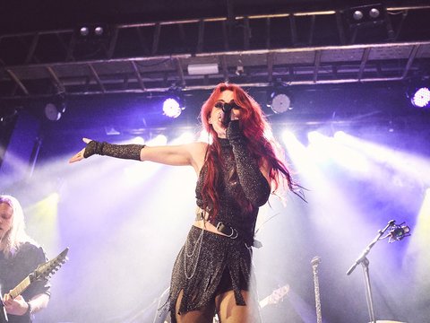 Eine Sängerin mit langen roten Haaren steht auf der Bühne und singt. Sie trägt ein schwarzes Outfit und hat einen energiegeladenen Auftritt. Im Hintergrund sind Musiker mit Gitarren zu sehen, während Bühnenlichter strahlen.