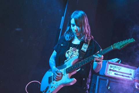 Eine Sängerin mit langen, brünetten Haaren spielt eine elektrische Gitarre auf der Bühne. Sie trägt ein schwarzes T-Shirt und eine Weste. Der Hintergrund ist dunkel und mit bunten Lichtern beleuchtet, während ein Verstärker im Hintergrund zu sehen ist.