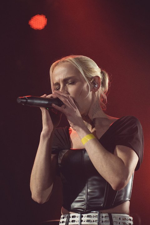 A female singer performs on stage, holding a microphone with both hands. She has short blonde hair and is dressed in a stylish outfit. The background features soft lighting, adding to the concert atmosphere. She appears focused and immersed in her performance.