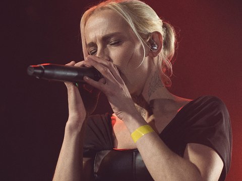 Eine Sängerin mit blonden Haaren trägt ein schwarzes Oberteil und hält ein Mikrofon. Sie scheint in einem musikalischen Moment vertieft zu sein. Der Hintergrund ist schwach beleuchtet, was eine konzentrierte Atmosphäre schafft.