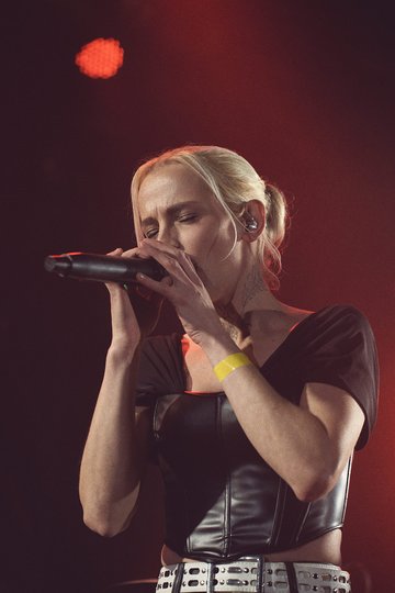 Eine Sängerin mit blonden Haaren trägt ein schwarzes Oberteil und hält ein Mikrofon. Sie scheint in einem musikalischen Moment vertieft zu sein. Der Hintergrund ist schwach beleuchtet, was eine konzentrierte Atmosphäre schafft.