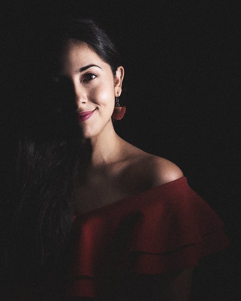 A woman with long, dark hair smiles softly in a dimly lit setting. She wears a red off-the-shoulder top and decorative earrings. Half of her face is illuminated, creating a dramatic contrast with the dark background.