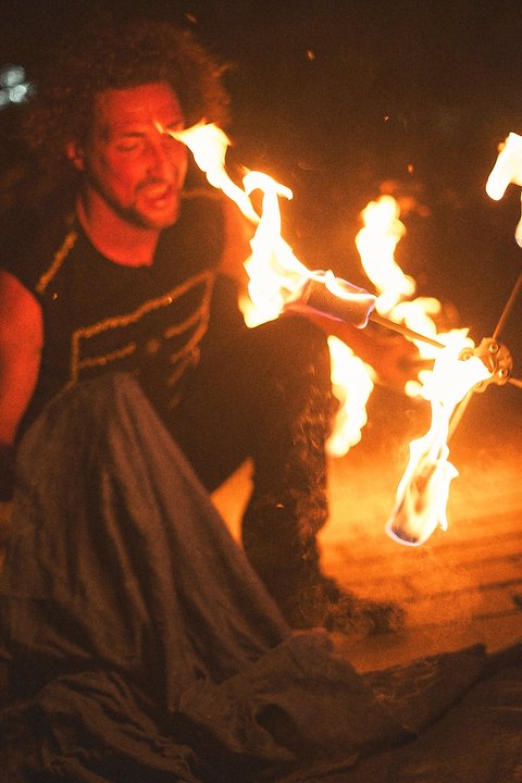 A performer kneels on the ground, skillfully manipulating flames with a fire staff. The bright orange flames contrast against the dark background, highlighting the intensity of the moment. The performer appears focused and engaged in the act of fire dancing.