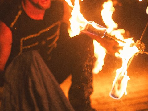 Ein Mann führt eine beeindruckende Feuershow auf, indem er brennende Fackeln in seinen Händen hält. Er lacht und scheint konzentriert zu sein, während die Flammen in der Dunkelheit leuchten. Die Atmosphäre ist aufregend und dynamisch.