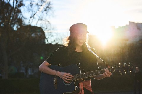 Eine junge Frau spielt Gitarre im Freien, während sie ins Sonnenlicht schaut. Sie trägt ein schwarzes T-Shirt und einen Hut. Die Atmosphäre ist warm und einladend, mit Sonnenschein im Hintergrund.