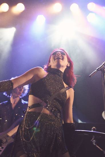 Eine Sängerin mit roten Haaren trägt ein glitzerndes, schwarzes Outfit und scheint glücklich auf der Bühne zu performen. Im Hintergrund ist ein Musiker zu sehen, während farbige Lichter den Raum erhellen.