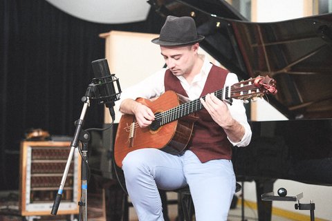Ein Musiker sitzt auf einem Stuhl und spielt Gitarre. Er trägt einen Hut und ein elegantes Oberteil. Neben ihm steht ein Mikrofon und im Hintergrund ist ein Klavier sichtbar. Die Atmosphäre wirkt künstlerisch und intim.