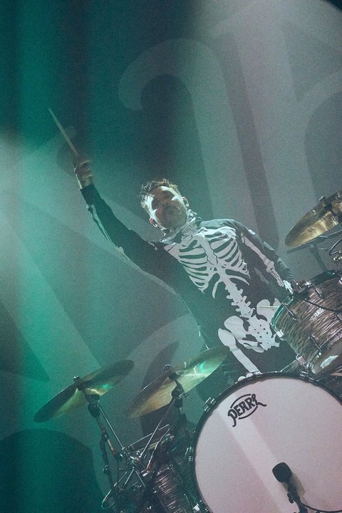 Ein Schlagzeuger in einem Kostüm mit einem Skelett-Muster hebt einen Drumstick in die Luft. Er steht hinter einem Schlagzeug und ist von leuchtenden Lichtern umgeben, die eine lebendige Konzertatmosphäre schaffen.
