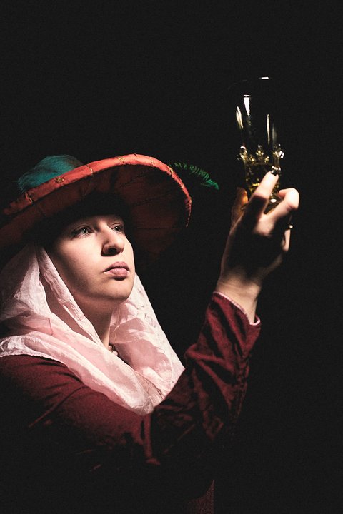 Eine Person trägt einen großen, bunten Hut und einen schlichten Schleier. Sie hält ein goldenes Objekt in der Hand und schaut es neugierig an. Der Hintergrund ist dunkel, wodurch die Person und das Objekt hervorgehoben werden.