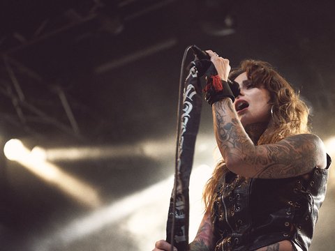 Eine Frau mit langen, lockigen Haaren singt leidenschaftlich in ein Mikrofon. Sie trägt ein schwarzes, ärmelloses Oberteil und hat sichtbare Tattoos an ihren Armen. Im Hintergrund sind Lichter und eine belebte Konzertatmosphäre zu erkennen.