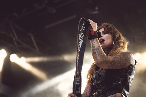 A tattooed woman passionately sings into a microphone on stage, wearing a black sleeveless outfit. Bright stage lights create a dramatic background, highlighting her intense performance.