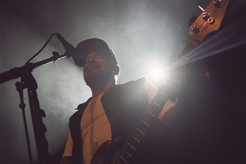 Ein Musiker steht auf einer Bühne, spielt Gitarre und spricht ins Mikrofon. Hinter ihm sind helle Lichter und Nebel zu sehen, die eine lebhafte Konzertatmosphäre schaffen. Der Fokus liegt auf dem Musiker und seiner Darbietung.