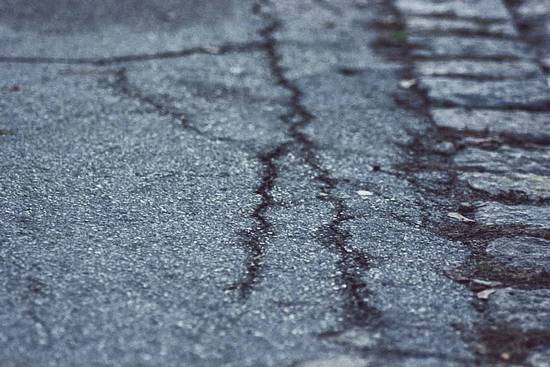 Die Nahaufnahme zeigt eine beschädigte Straße mit Rissen und unebenen Stellen. Der Asphalt hat eine graue Farbe und weist Spuren von Abnutzung auf. An der Seite sind unregelmäßige Steinplatten sichtbar.