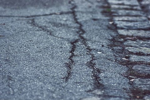 Die Nahaufnahme zeigt eine beschädigte Straße mit Rissen und unebenen Stellen. Der Asphalt hat eine graue Farbe und weist Spuren von Abnutzung auf. An der Seite sind unregelmäßige Steinplatten sichtbar.