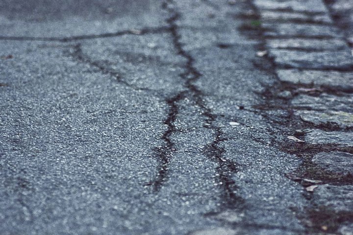 Die Nahaufnahme zeigt eine beschädigte Straße mit Rissen und unebenen Stellen. Der Asphalt hat eine graue Farbe und weist Spuren von Abnutzung auf. An der Seite sind unregelmäßige Steinplatten sichtbar.