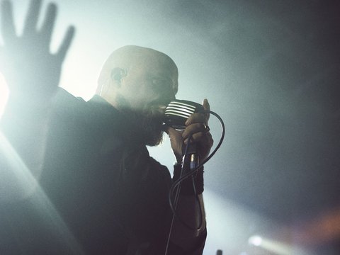 Ein Sänger steht auf der Bühne und hält ein Mikrofon in der Hand. Er hat einen rasierten Kopf und trägt dunkle Kleidung. Sein Gesicht ist in Gegenlicht getaucht, während er mit ausgestreckter Hand zum Publikum spricht oder singt. Die Atmosphäre wirkt dynamisch und kraftvoll.