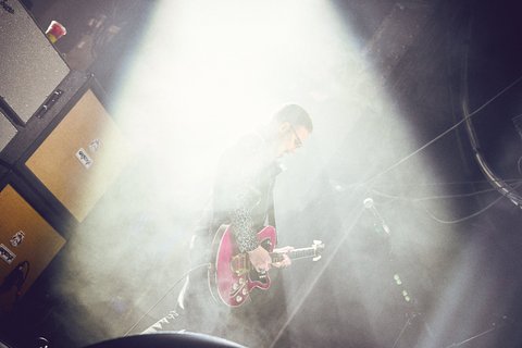 Ein Musiker steht auf der Bühne und spielt eine pinke Gitarre. Er ist von Nebel umgeben, während Scheinwerferlicht auf ihn scheint. Im Hintergrund sind Verstärker sichtbar. Die Atmosphäre ist intensiv und lebhaft, passend zu einem Konzert.