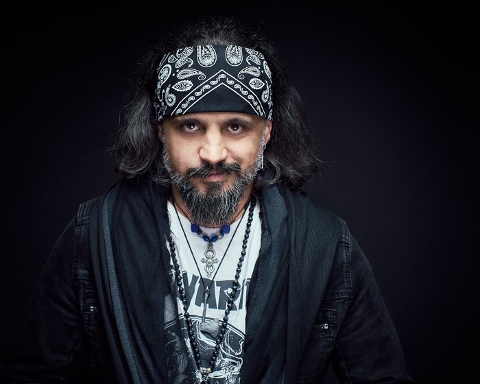 Ein Mann mit langen, dunklen Haaren und einem schwarz-weißen Bandana steht vor einem dunklen Hintergrund. Er trägt ein schwarzes Oberteil und mehrere Ketten. Sein Gesicht zeigt einen ernsten Ausdruck.