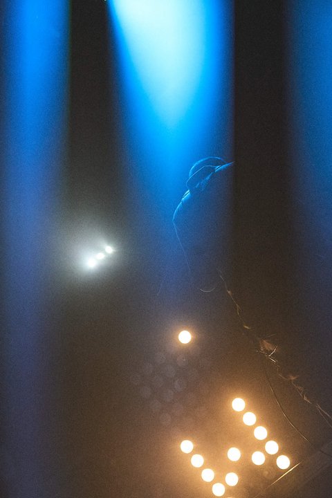 Ein silhouettierter Künstler steht im Vordergrund, umgeben von bunten Lichtstrahlen, die von oben abstrahlen. Die Bühne ist schwach beleuchtet und es sind einige glänzende Scheinwerfer zu erkennen, die eine stimmungsvolle Atmosphäre schaffen.