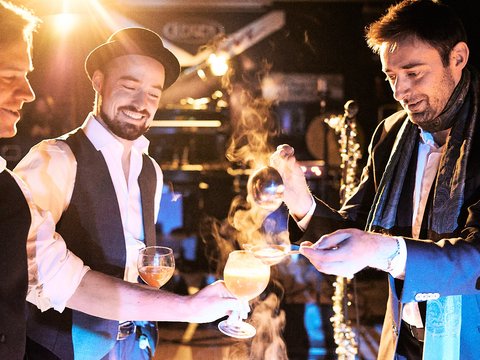Drei Männer in formeller Kleidung stehen in einer feierlichen Atmosphäre. Einer von ihnen hält ein glas mit einem rauchenden Cocktail, während die anderen interessiert zuschauen. Sie scheinen bei einer besonderen Veranstaltung oder Feier zu sein.