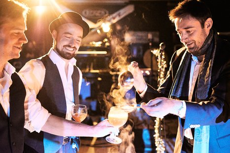 Drei Männer in formeller Kleidung stehen in einer feierlichen Atmosphäre. Einer von ihnen hält ein glas mit einem rauchenden Cocktail, während die anderen interessiert zuschauen. Sie scheinen bei einer besonderen Veranstaltung oder Feier zu sein.