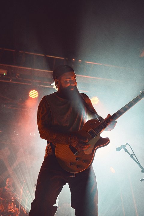 Ein Musiker spielt leidenschaftlich Gitarre auf einer Bühne, umgeben von bunten Lichtern und Nebel. Er trägt einen Hut und hat einen Bart, während er sich intensiv auf sein Spiel konzentriert. Die Atmosphäre ist energetisch und musikalisch.