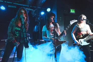 Eine Rockband aus drei Musikerinnen steht auf der Bühne, umgeben von bläulichem Nebel. Eine Sängerin mit Mikrofon, eine Gitarristin und eine Bassistin spielen leidenschaftlich. Im Hintergrund sind Lichter zu sehen, die die energische Atmosphäre des Konzerts verstärken.