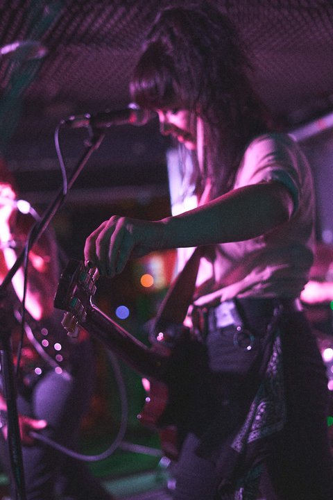 Eine Musikerin auf der Bühne spielt Gitarre. Sie trägt ein lässiges Outfit und konzentriert sich auf ihre Performance. Im Hintergrund sind unscharfe Silhouetten anderer Bandmitglieder zu erkennen. Die Beleuchtung ist schwach, mit einem sanften, bunten Glühen.