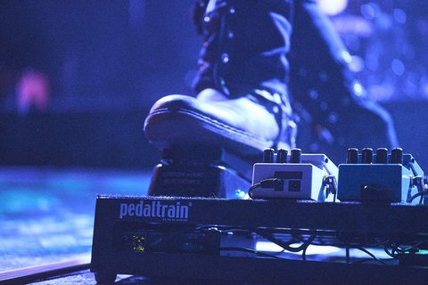 Ein Fuß in einem schwarzen Stiefel drückt auf ein Effektpedal auf der Bühne. Neben dem Pedal sind mehrere weitere Effektgeräte sichtbar. Die Szene vermittelt die Atmosphäre eines Live-Konzerts.