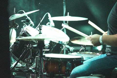 Ein Schlagzeuger spielt leidenschaftlich auf seinem Schlagzeug. Sticks treffen auf die Trommeln, während der Musiker in Jeans und T-Shirt sichtbar ist. Licht und Dämpfung erzeugen eine energiegeladene Atmosphäre.