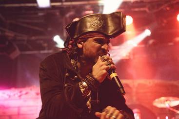 Ein Sänger in einem Piratenkostüm hält ein Mikrofon und performt auf einer Bühne. Er trägt einen auffälligen Hut und hat mehrere Schmuckstücke an. Die Bühne ist mit bunten Lichtern beleuchtet, die eine lebhafte Atmosphäre schaffen.