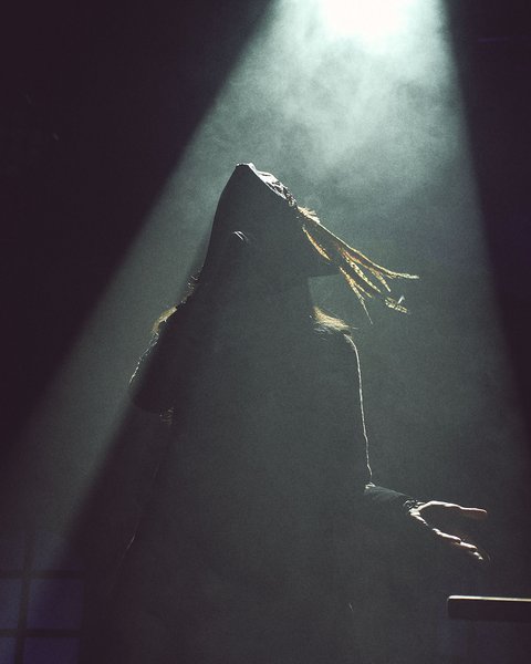 Eine silhouettenhafte Figur mit langen, schwingenden Haaren steht im Scheinwerferlicht. Umgeben von Nebel, hebt sie die Hände und wirkt Ausdruck von Emotion und Drama. Der Hintergrund ist dunkel, was die zentrale Figur betont.