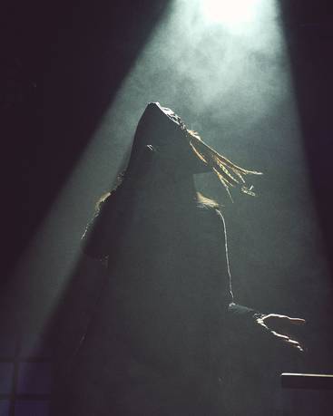 Eine silhouettenhafte Figur mit langen, schwingenden Haaren steht im Scheinwerferlicht. Umgeben von Nebel, hebt sie die Hände und wirkt Ausdruck von Emotion und Drama. Der Hintergrund ist dunkel, was die zentrale Figur betont.