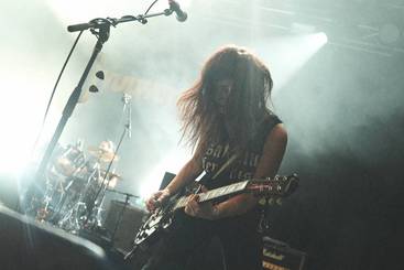 Eine Musikerin spielt Gitarre auf der Bühne. Ihr lange, lockige Haare wehen im Rhythmus der Musik. Im Hintergrund ist ein Schlagzeuger zu sehen. Das Licht ist gedämpft, und die Atmosphäre wirkt intensiv und energisch.