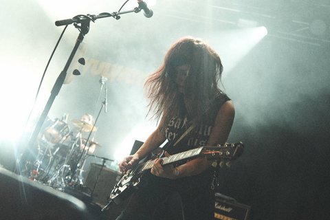 Eine Musikerin spielt Gitarre auf der Bühne. Ihr lange, lockige Haare wehen im Rhythmus der Musik. Im Hintergrund ist ein Schlagzeuger zu sehen. Das Licht ist gedämpft, und die Atmosphäre wirkt intensiv und energisch.