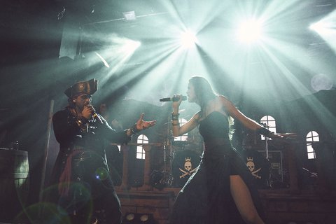 Zwei Musiker treten auf einer Bühne auf. Einer trägt ein Kostüm und hält ein Mikrofon, während die Sängerin in einem eleganten, schwarzen Kleid singt. Helle Scheinwerfer strahlen über die Bühne und schaffen eine dramatische Atmosphäre.