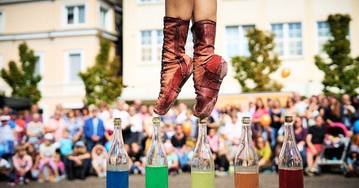 A performer in brown boots balances on one foot above five colorful bottles filled with liquids. The background shows a crowd watching the performance, creating a lively atmosphere. The focus is on the skillful balancing act near the bottles.