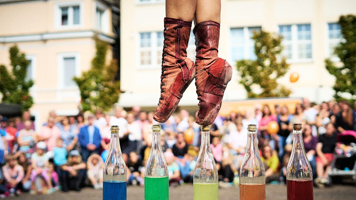 Eine Person in speziellen Tanzschuhen steht auf der Spitze von zwei angedeuteten, bunten Flaschen. Im Hintergrund ist eine Menschenmenge zu sehen, die aufmerksam zuschaut. Die Szenerie vermittelt eine festliche Atmosphäre mit einem Performance-Aspekt.