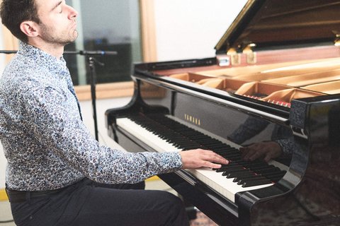 Ein Pianist sitzt konzentriert an einem schwarzen Flügel und spielt. Er trägt ein gemustertes Hemd und blickt mit geschlossenen Augen in die Höhe, während seine Hände über die Tasten gleiten. Die Umgebung ist hell erleuchtet und trägt zur musikalischen Atmosphäre bei.
