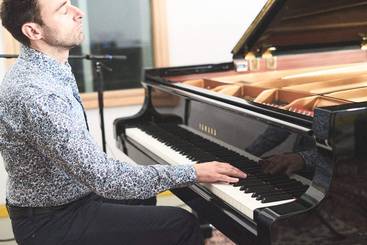 Ein Pianist sitzt konzentriert an einem schwarzen Flügel und spielt. Er trägt ein gemustertes Hemd und blickt mit geschlossenen Augen in die Höhe, während seine Hände über die Tasten gleiten. Die Umgebung ist hell erleuchtet und trägt zur musikalischen Atmosphäre bei.